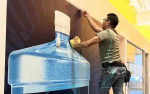 Installing large format wallpaper featuring a water bottle graphic, with a technician smoothing out the design, demonstrating 360CSG's expertise in wall graphics.